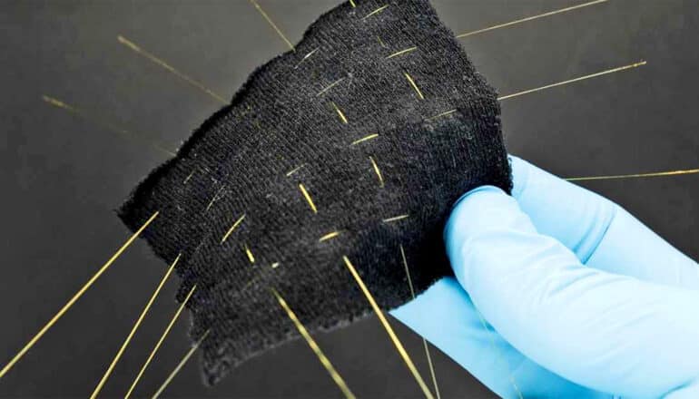 A researcher wearing a blue glove holds a small black square of fabric that has thin glass fibers woven through it.