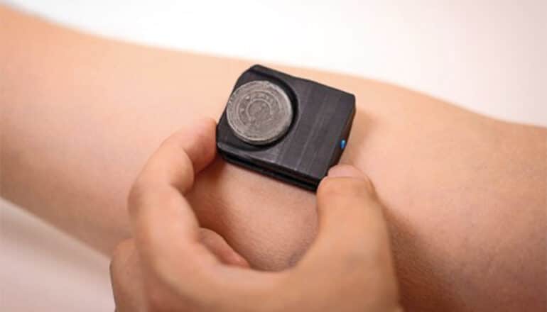 A person holds the wearable device on the skin on their arm. The device is a small black rectangle with a gray circular piece on top.