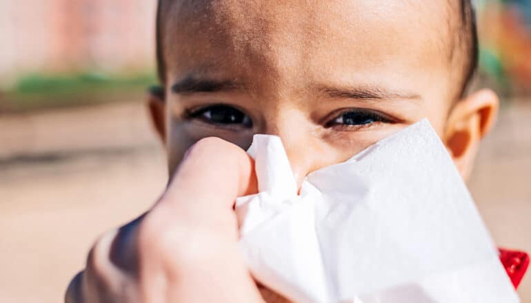 An adult puts a tissue up to a child's nose as the kid grimaces and squints.