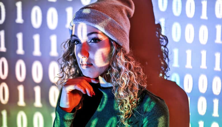 A young woman wearing a beanie puts her hand to her chin while binary code is projected on her face and the wall behind her.