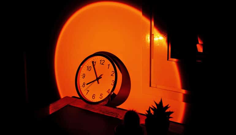 A clock sits on a shelf against a wall as a bright orange light illuminates it.