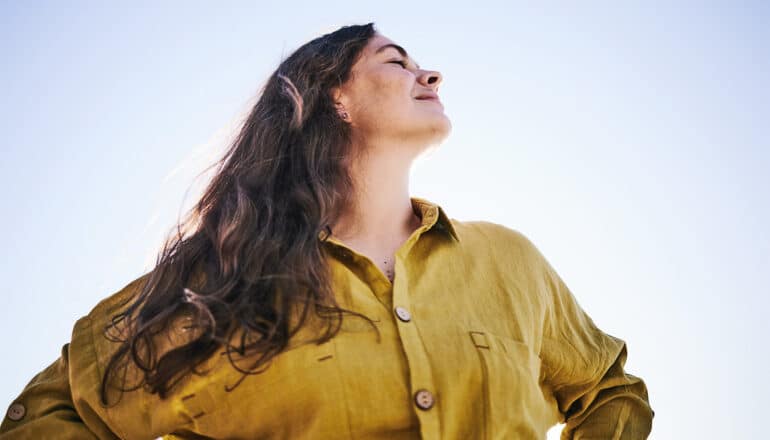 A woman tilts her head back, smiles, and closes her eyes as she puts her hands on her hips.