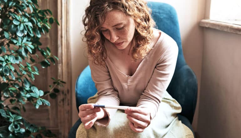 A woman looks down at a pregnancy test while sitting in a chair next to a window.