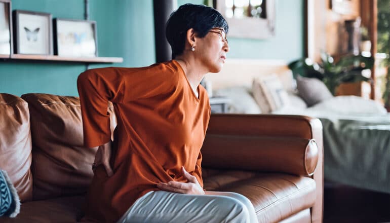 A woman holds her lower back as she gets up from a couch grimacing in pain.