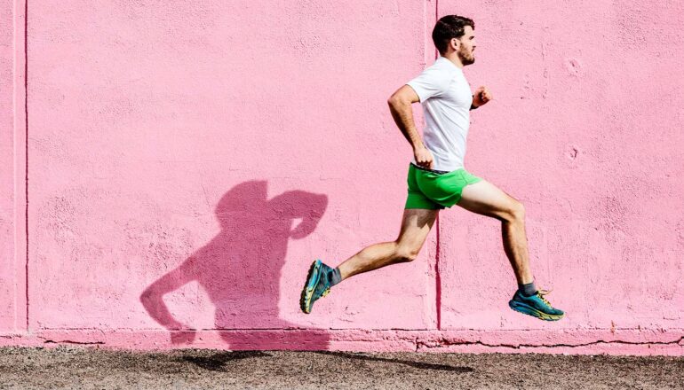 A man wearing a white shirt, green shorts, and green running shoes runs past a pink wall.