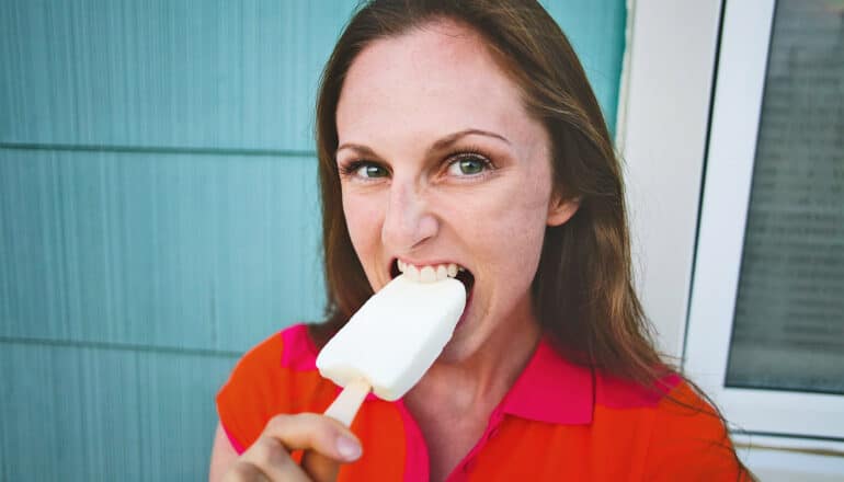 A woman in a red shirt takes a big bite of a white ice cream bar while standing in front of a blue wall.