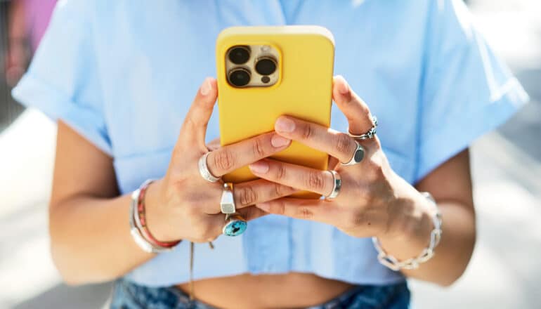 A young woman wearing many rings holds her yellow phone in both hands in front of her.