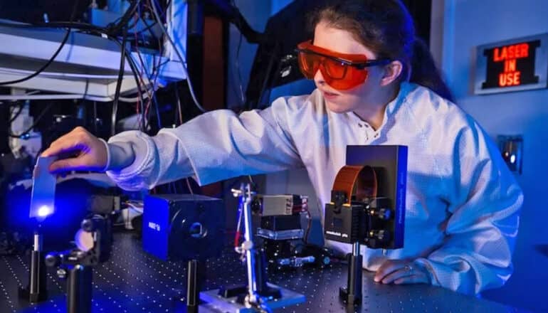 A researcher wearing a white lab coat and red safety goggles uses a laser device in a lab.