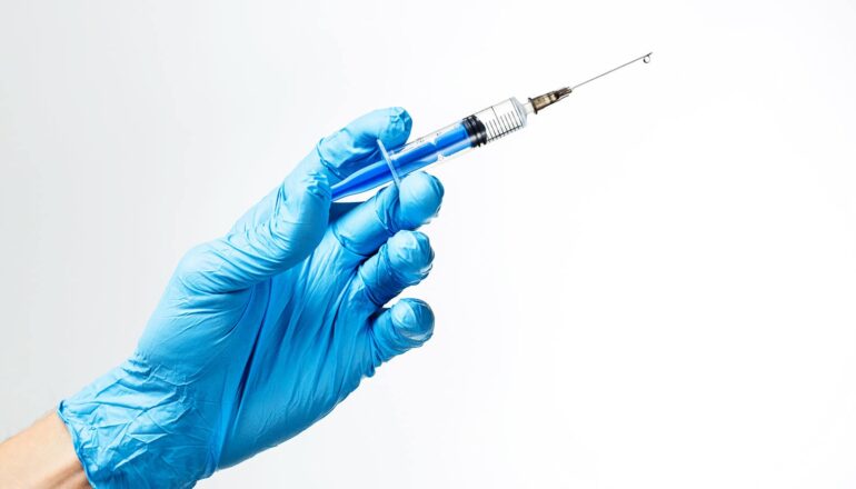 A blue-gloved hand holds a vaccine syringe in front of a white background.