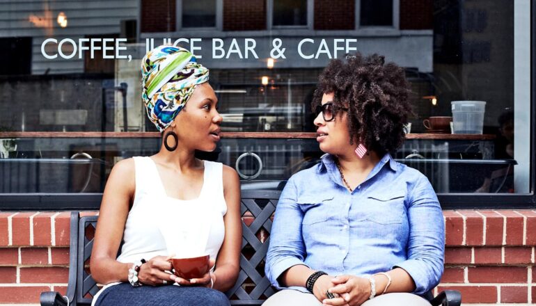 Two friends talk while sitting on a bench outside a coffee shop.