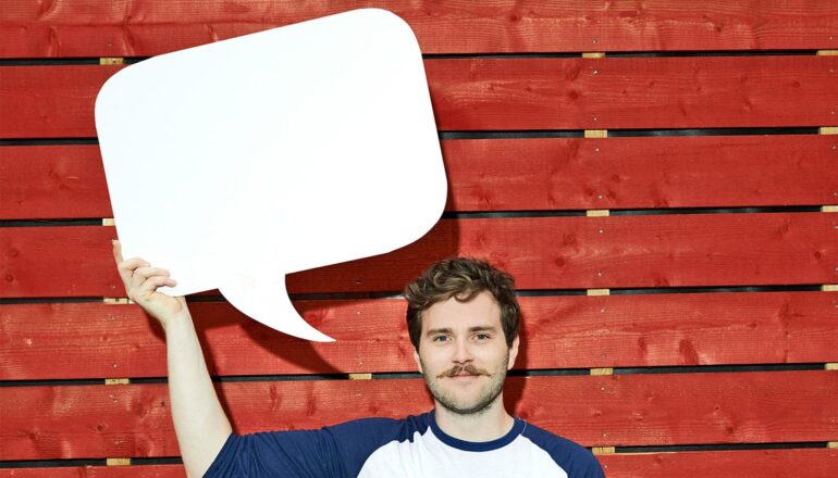 A man holds up a white cardboard cutout of a speech bubble as he stands against a red wood wall.