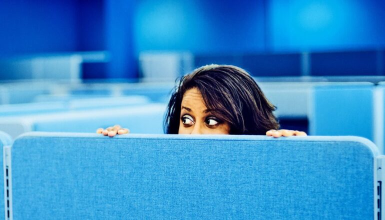 An office worker peeks over a bright blue cubicle wall and looks surprised.