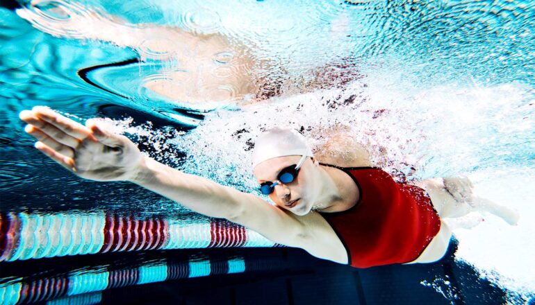 A young woman swims underwater in an athletic pool while wearing a cap and goggles.