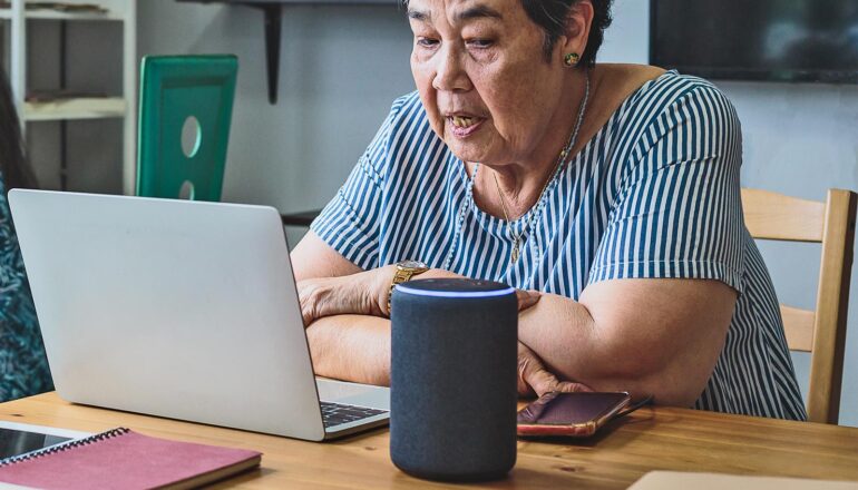 A woman sits at a table with her laptop, voice assistant device, and cell phone.