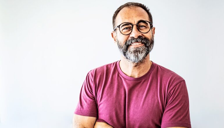 A middle age man in a red shirt and glasses smiles while standing in front of a white background.