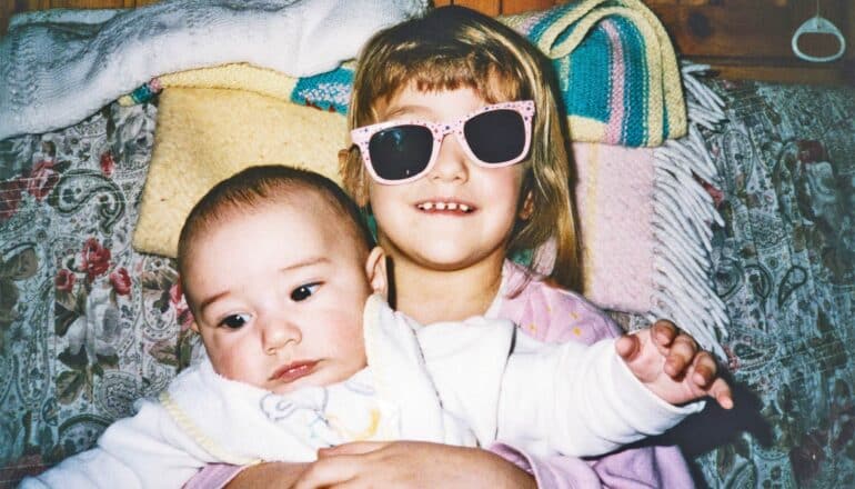 A young girl wearing askew sunglasses smiles for a picture as she holds her baby sibling.