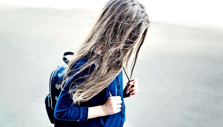 A young girl looks downward while walking to school wearing a backpack.