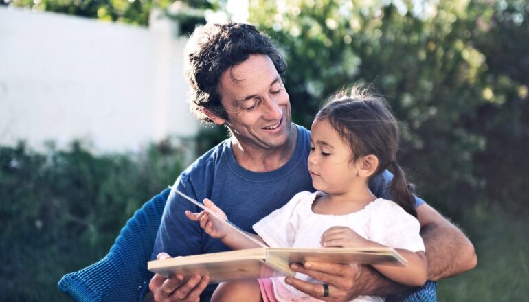 A father reads to his daughter while sitting outside.