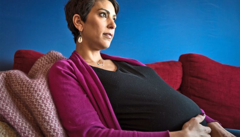 A pregnant woman sits on a couch and looks off into the distance.