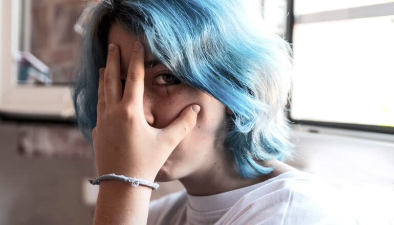 A teen with blue hair holds a hand up to her face while looking at the camera.