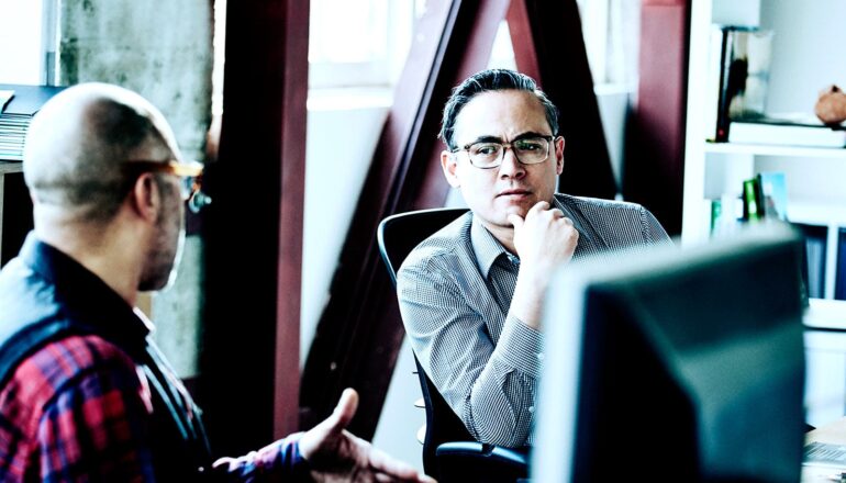 An employee speaks to a manager in an office as he looks back with a skeptical expression.