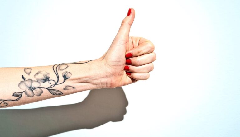 A woman with red nails and a flowery tattoo on her arm holds up a thumbs up sign.