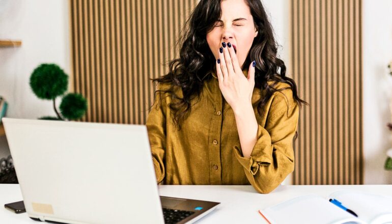 A young woman working at a laptop covers her mouth and closes her eyes as she yawns from boredom.