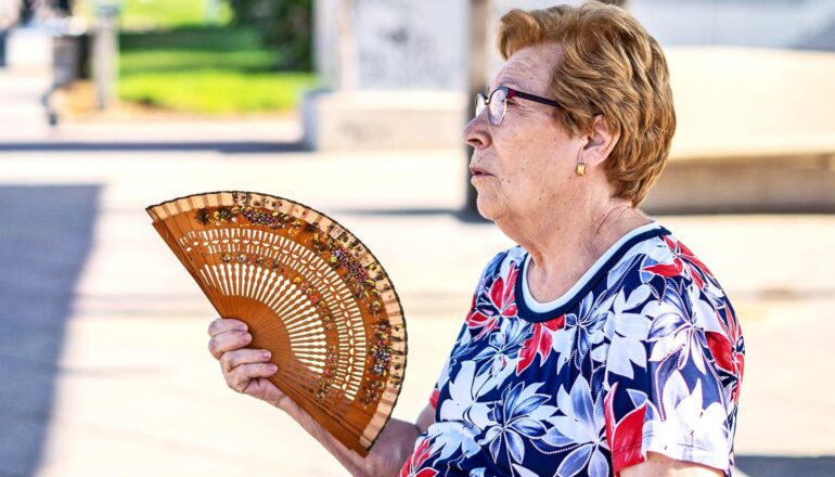 An older woman sits outside while fanning herself with a large fan.