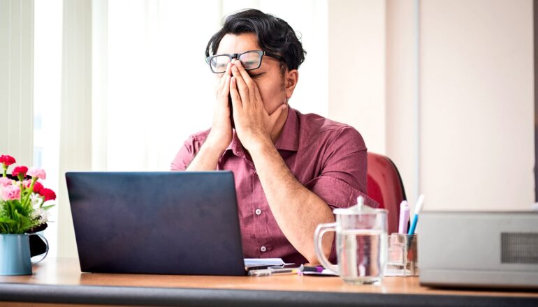 An employee puts both hands over their face under their glasses as they sit in front of a laptop at a desk.