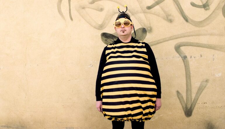 A man dressed in a bee costume and sunglasses stands against a beige wall.