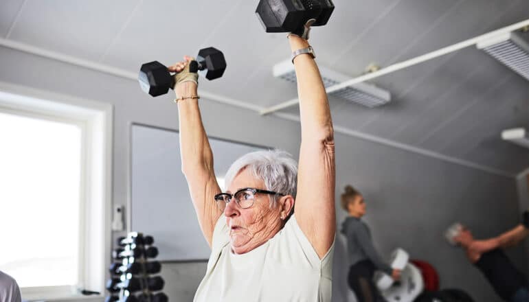 An older woman holds two free weights high over her head in a gym.