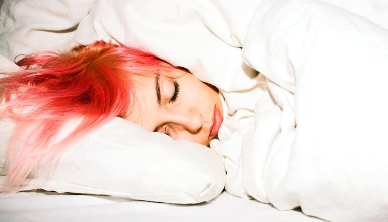 A young woman with dyed red hair sleeps in a bed with white sheets and blankets.