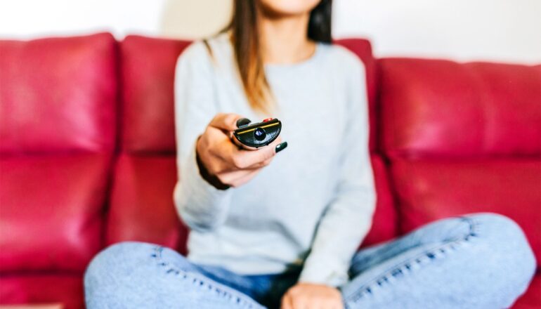 A young woman points a remote at a tv while sitting on a red couch.
