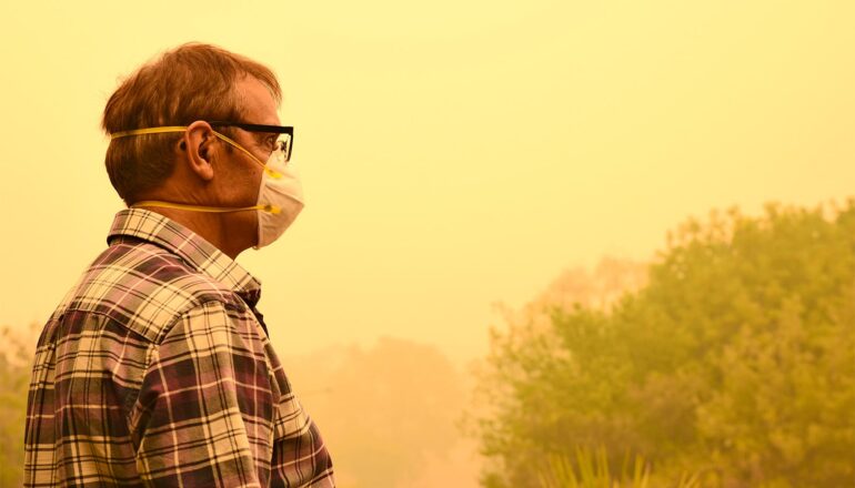 A man stands outside wearing a face mask while surrounded by yellow wildfire smoke in the air.