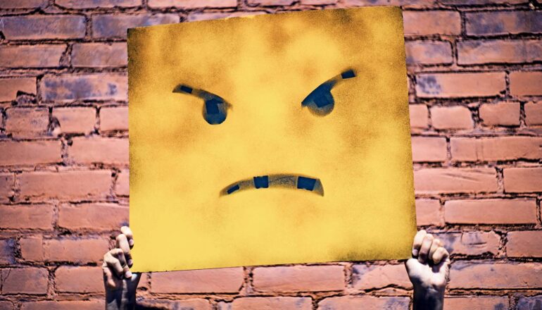 A person holds up a yellow piece of cardboard with an angry face drawn on it.