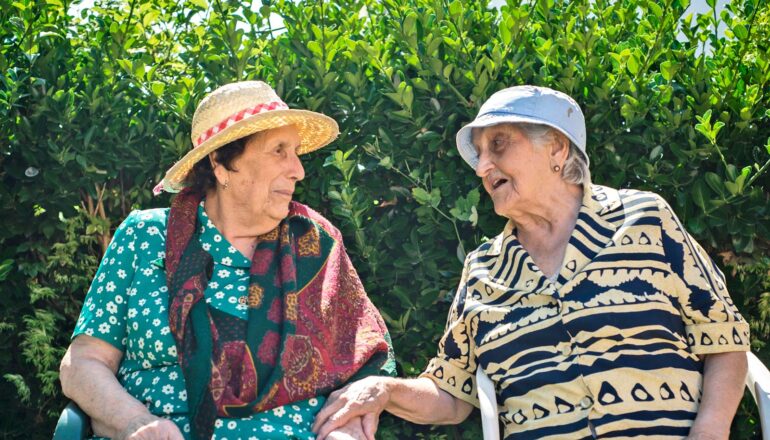 Two older women chat while sitting outside in front of a green hedge.