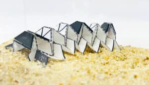 An origami robot made of what looks like folded cardboard stands on sand in a small enclosure.