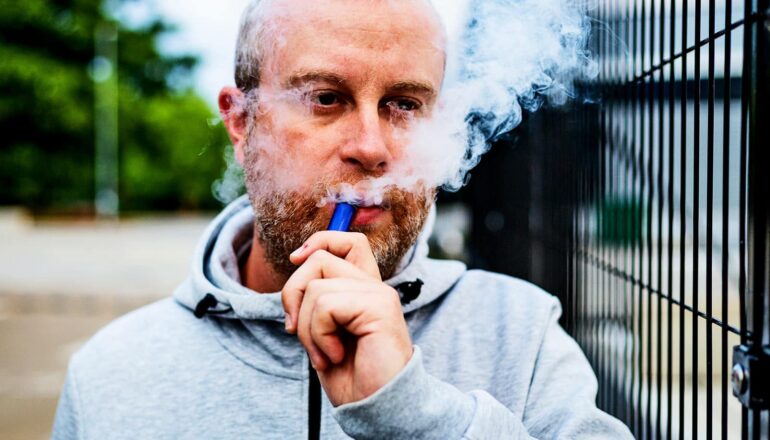 A man vaping as he walks past a fence outside.