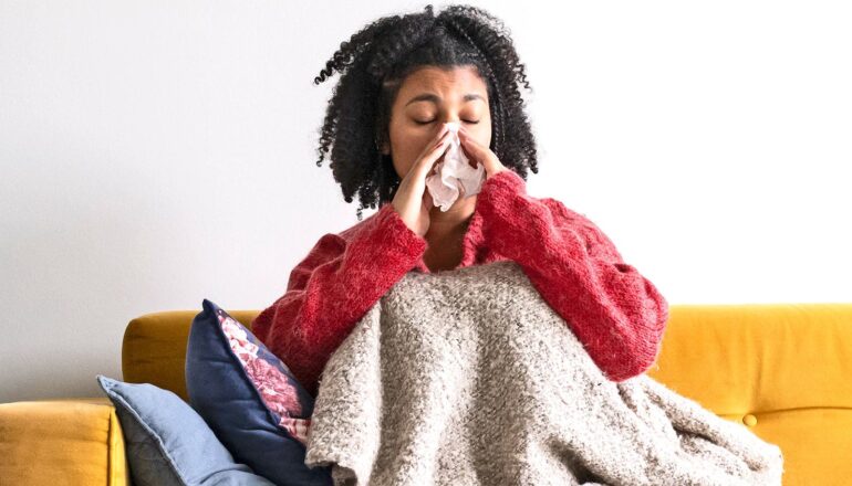 A woman blows her nose while sitting on a couch under a blanket.