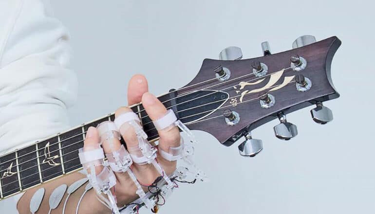 A person wears a gadget with wires and electronics on their hand while playing guitar.