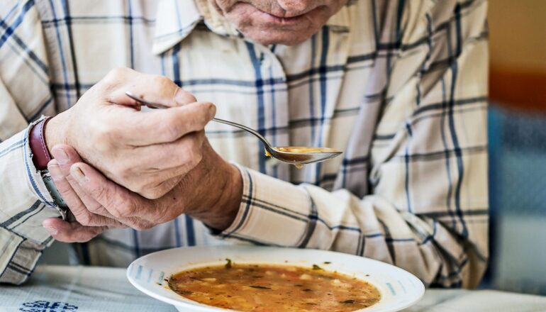 A man eats soup with a spoon in one hand and the other hand steadying it.