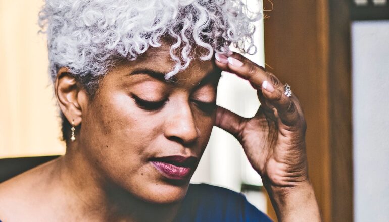 A woman with gray hair touches her temple and closes her eyes.