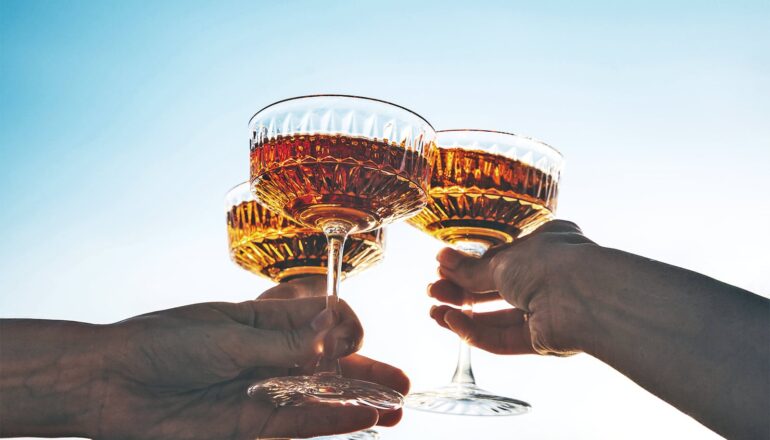 People clink glasses filled with dark liquid in front of a blue sky.