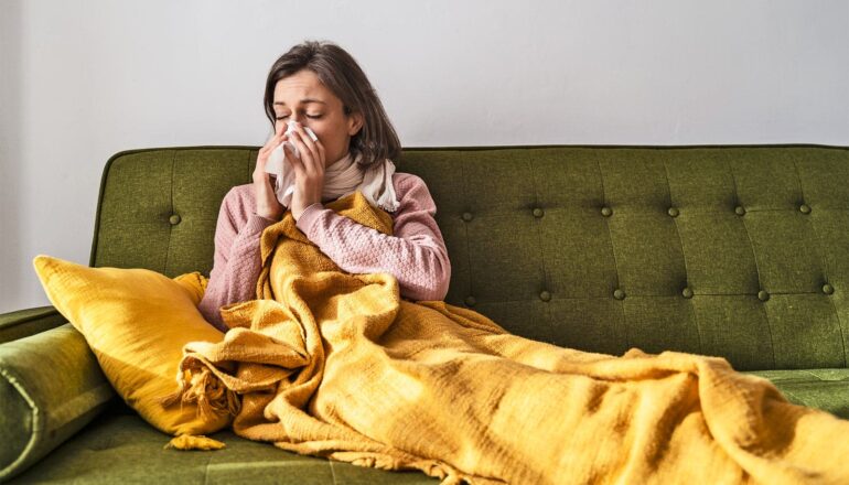 A woman blows her nose while sitting on a couch under a blanket.