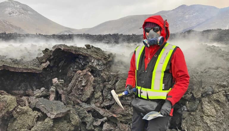 A researcher in protective gear stands in front of steaming lava rock.
