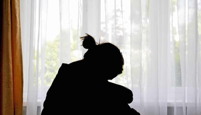 A person sits on a bed with their head on their knees in silhouette.