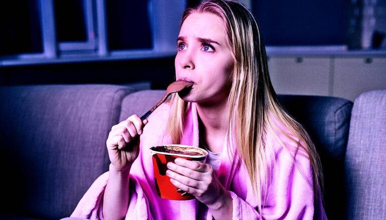 A woman eats ice cream while watching a movie on her couch.