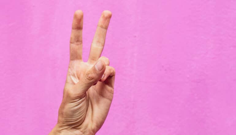 A person holds up two fingers in front of a pink background.