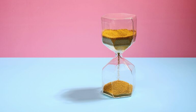 An hourglass with sand running to the bottom sits on a blue surface in front of a pink background.