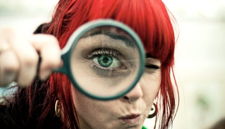 A woman with dyed red hair looks through a magnifying glass.